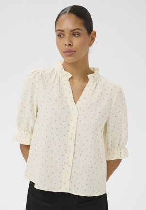 Femme portant un chemisier crème à petit imprimé floral, col à volants et manches bouffantes mi-longues, debout devant un fond uni.