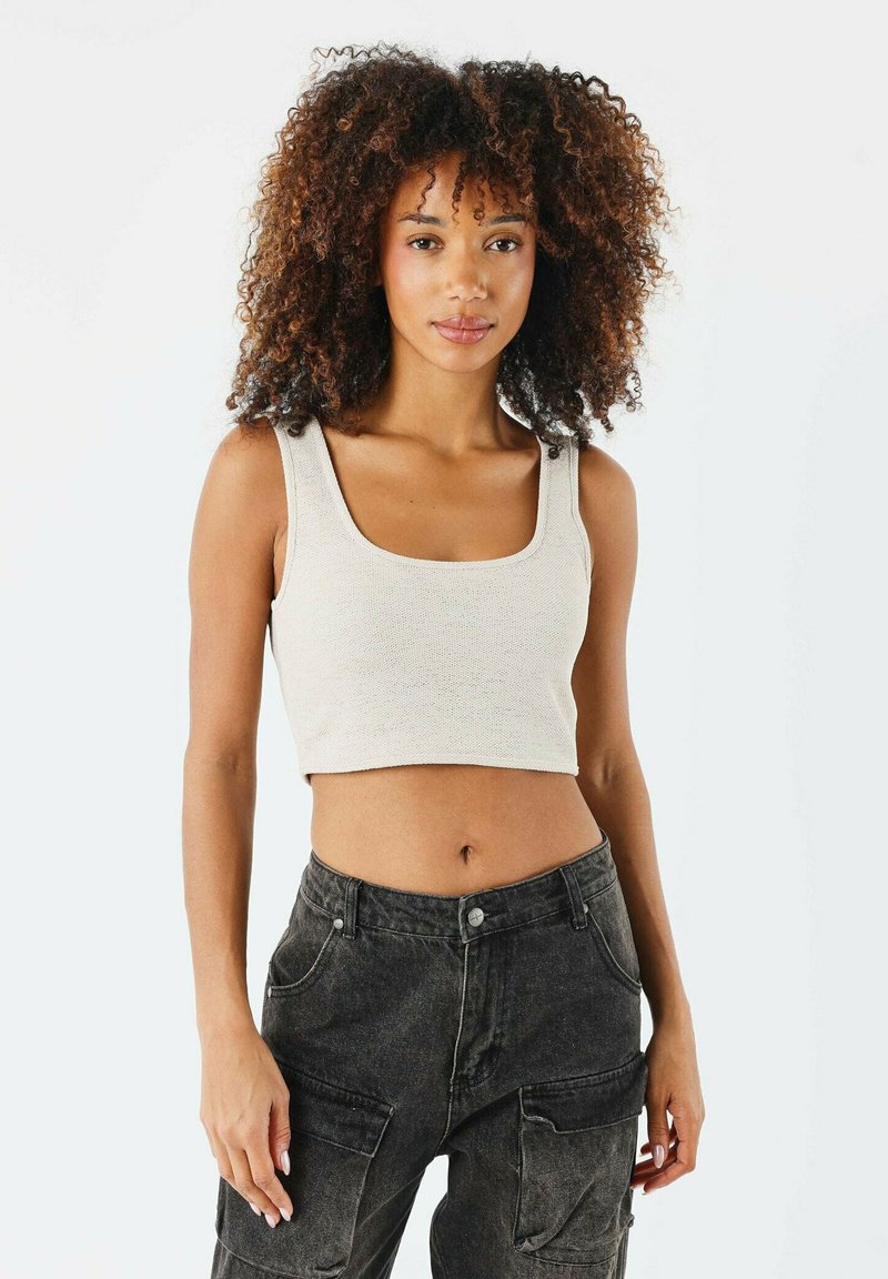 Haut cropped beige avec un large décolleté carré et des bretelles épaisses, associé à un pantalon cargo sombre avec des poches surdimensionnées.