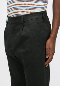 Homme portant un pantalon sur mesure gris foncé et un haut en tricot à rayures multicolores, debout avec la main droite détendue le long du corps.