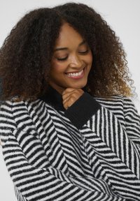 Sieviete ar cirtainiem matiem smaida, ģērbusies melnbaltā svītrainā mīkstā džemperī ar melniem piedurknēm un apkakli.