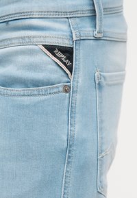 Φωτεινό γαλάζιο τζιν τσέπης που δείχνει μια μαύρη τριγωνική ετικέτα της μάρκας Replay ραμμένη μέσα στην μπροστινή δεξιά τσέπη.