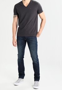 Mörkgrå v-ringad t-shirt, slim fit, bärs med mörkblå jeans och ljusgrå sneakers. T-shirten har en liten logga på bröstet.