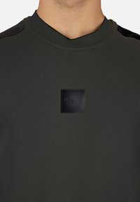 Sudadera verde oliva con cuello en V, que presenta un parche de logo negro texturizado en el pecho y detalles negros en los hombros. Textura de tela suave.
