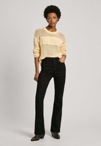Suéter amarillo de punto con trama abierta y detalles de canalé, combinado con jeans negros de cintura alta y acampanados, y botas marrones de tacón.