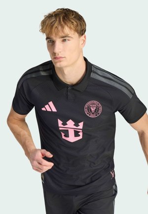 Young man in black and pink Inter Miami soccer jersey running forward against plain light background.