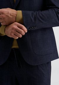 Marineblauer kariert Blazer mit Revers und vierknöpfigem Ärmeldetail, kombiniert mit einem senfgelben Strickpullover darunter.