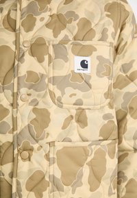 Quilted beige camouflage jacket with a chest pocket featuring a logo patch, round buttons, and a smooth, textured fabric finish.