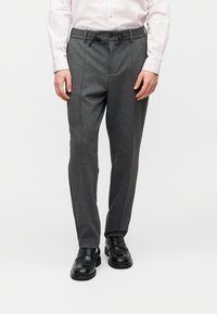 Άνδρας που φοράει γκρι παντελόνι με κορδόνι στη μέση και μαύρα δερμάτινα παπούτσια χωρίς κορδόνια, στέκεται μπροστά σε απλό λευκό φόντο.