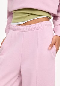 Světlé růžové tepláky s elastickým pasem, mají boční kapsy a švy. Pod nimi je vrstvený zelený top.