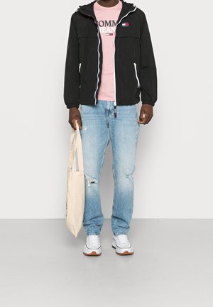 Giacca leggera nera con zip bianche, T-shirt rosa con testo, jeans chiari strappati, sneaker bianche e una borsa a tracolla crema.
