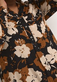 Chemise à motif floral marron et beige avec une patte de boutonnage noire portée par une personne levant un bras.