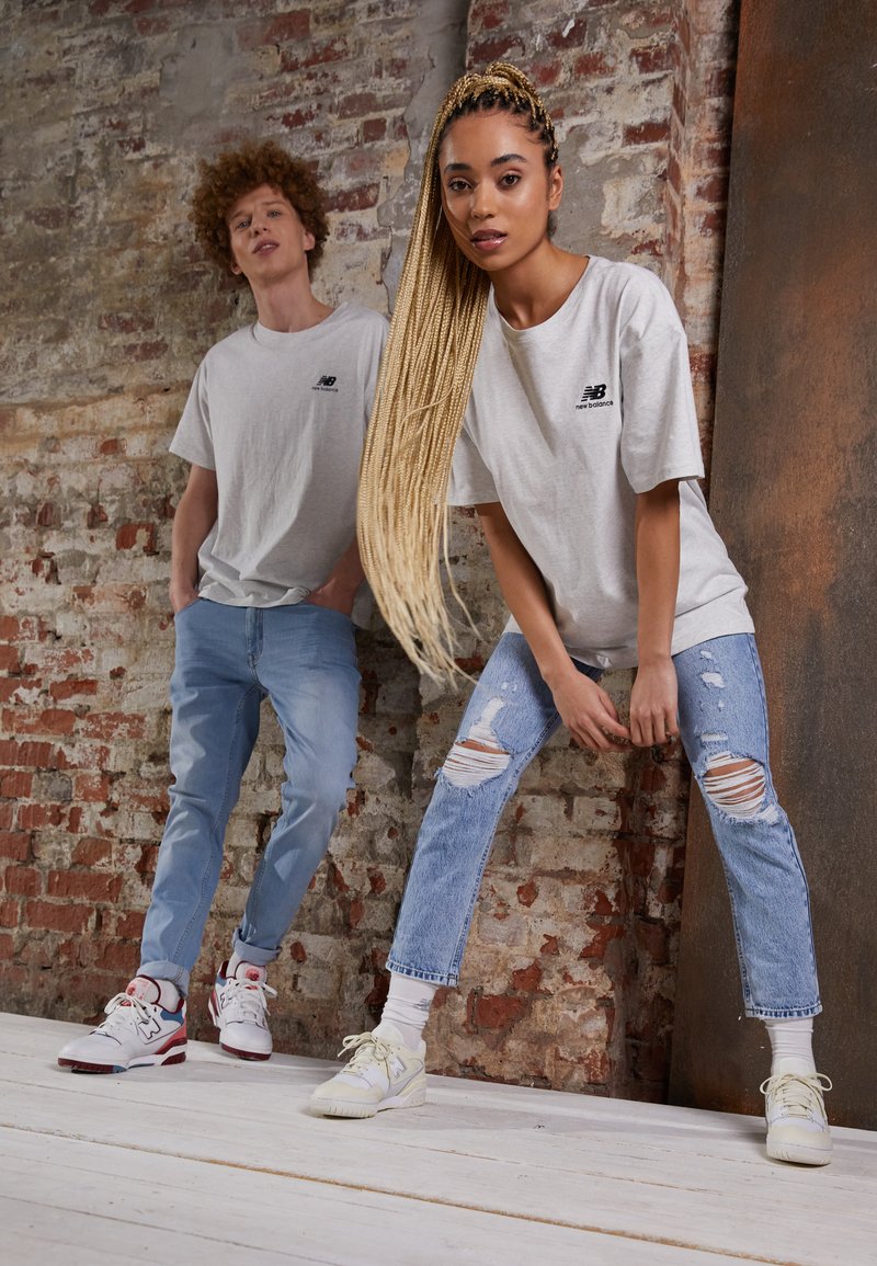 Due modelli posano contro un muro di mattoni. Indossano t-shirt grigio chiaro, jeans in denim strappati e sneakers. Caratteristiche distintive includono lunghe trecce.