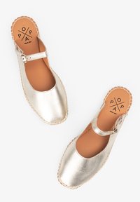 Zapatos de cuero metálico dorado con diseño de punta cerrada, correa en el tobillo y suela de goma cosida. La plantilla presenta un logo de la marca.