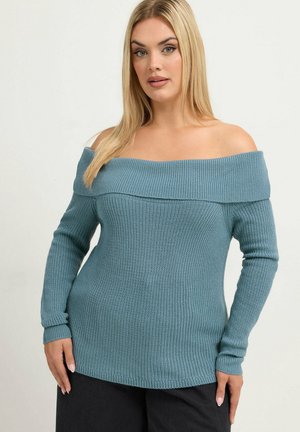 Femme aux longs cheveux blonds portant un pull tricoté bleu clair à décolleté bardot et un pantalon noir, posant sur un fond uni.