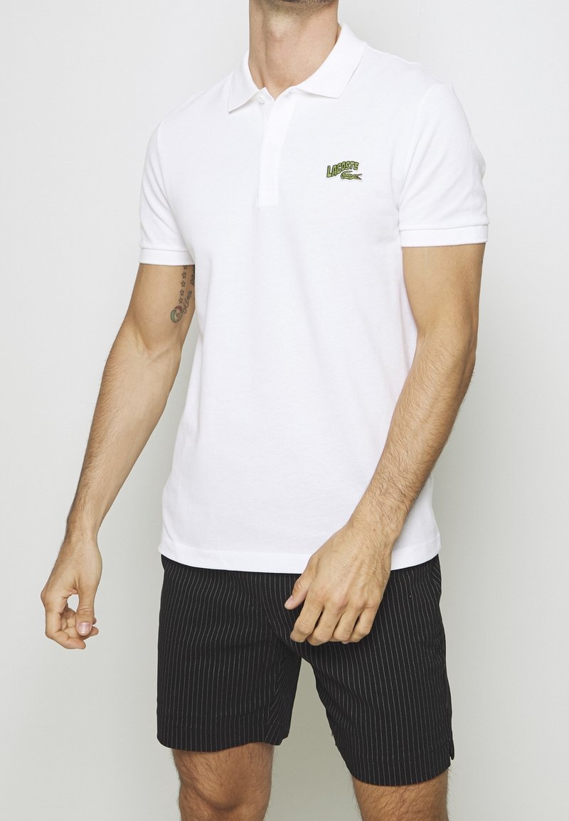 Homme portant un polo blanc Lacoste à manches courtes et un short noir à fines rayures, debout devant un fond clair uni.