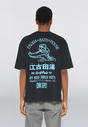 Person trägt ein schwarzes T-Shirt mit blauer Grafik und Text, das eine Cartoon-Krokodilfigur, japanische Schriftzeichen und Kontaktinformationen auf der Rückseite zeigt.