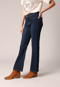 Jean évasé bleu foncé avec une texture lisse, doté de poches avant et d'une fermeture à bouton, porté avec des bottines à talons en daim marron.