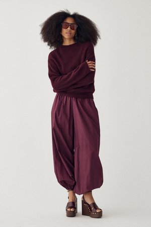 Nagy göndör hajjal rendelkező nő sötét napszemüvegben, bordó pulóverben, széles, magas derekú nadrágban és barna talpú szandálban.