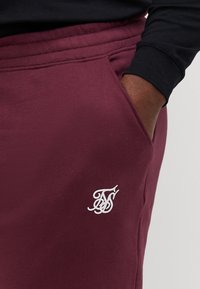 Joggers burdeos de tela suave, con un bolsillo lateral y un logo bordado en blanco en el muslo. Cintura elástica para mayor comodidad.