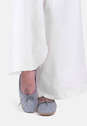 Fødder iført lysegrå ruskind balletflade sko med sløjfedetalje, delvist dækket af lange hvide bukser med vide ben.