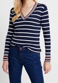Marineblauer Pullover mit weißen horizontalen Streifen, V-Ausschnitt und gerippter Struktur. Rote Akzente betonen die Bündchen und den Kragen.