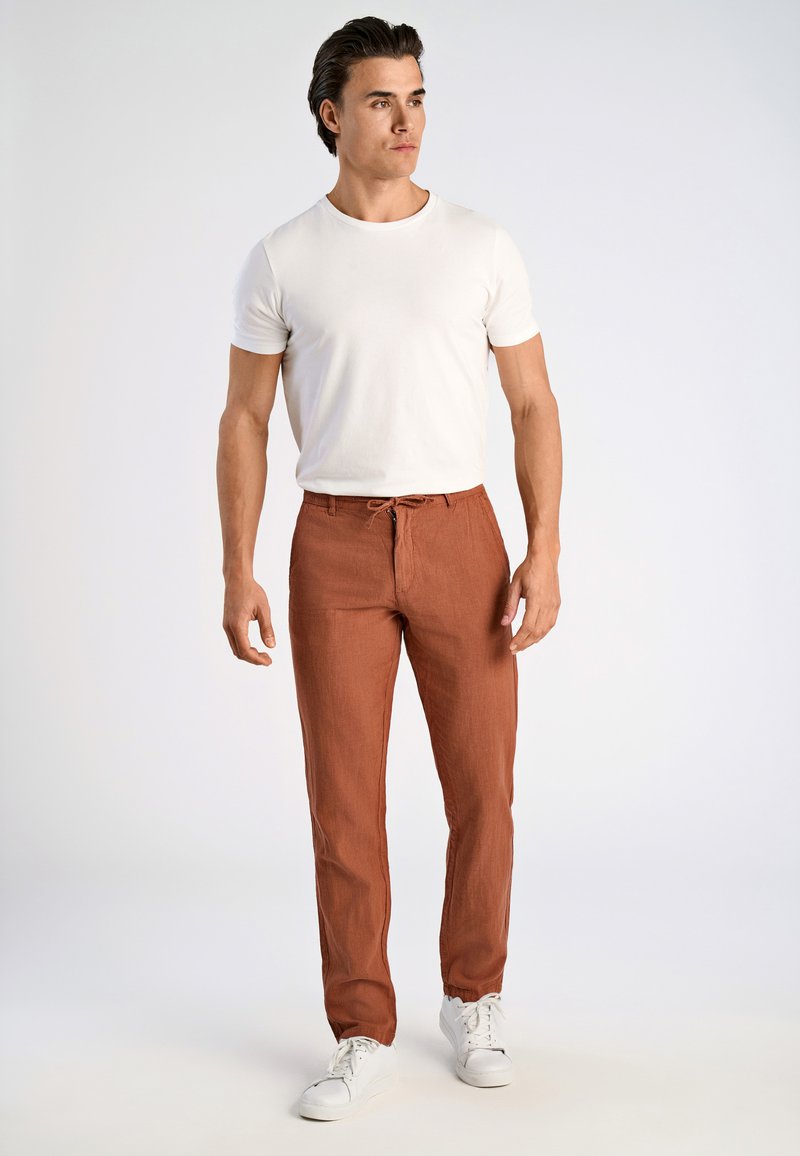 Roestkleurige linnen broek met een trekbare tailleband, gecombineerd met een getailleerd wit T-shirt en witte sneakers. Model staat in een neutrale omgeving.