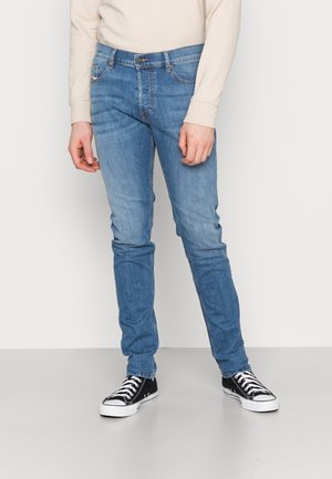 Personne portant un jean slim bleu clair, des baskets noires et blanches, et une chemise crème à manches longues, debout devant un fond uni.