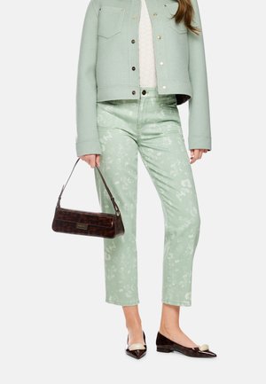 Frau trägt hellgrüne Cropped-Jacke und grüne Hose mit Blumenmuster, hält kleine braune Handtasche mit dunklen Absätzen.