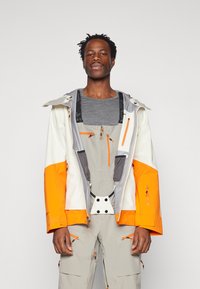 Ski jacket in white, orange, and gray with multiple zippered pockets, adjustable cuffs, and a detachable hood, worn over gray outfit.
