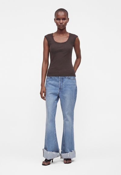 Femme portant un haut sans manches marron foncé ajusté, un jean large bleu clair à revers, et des talons ouverts noirs, debout devant un fond uni.