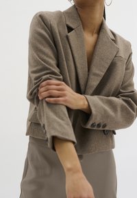 Blazer a manica corta marrone realizzato in tessuto testurizzato, con rever a punta, tasca frontale e polsini con bottoni. Abbinato a una gonna beige.