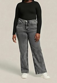 Personne portant un haut noir à manches longues, un jean gris délavé taille haute et des chaussures blanches slip-on, debout contre un fond clair uni.