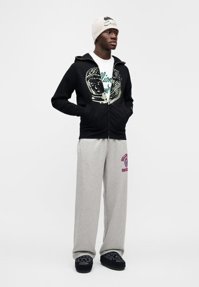 Μαύρο φούτερ με φερμουάρ και γραφικό σχέδιο, λευκή μπλούζα, γκρι παντελόνι φόρμας με λογότυπο, μαύρες παντόφλες και ανοιχτόχρωμο σκουφάκι.