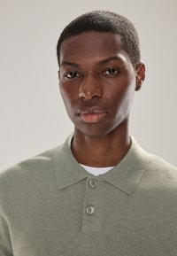 Kaki groene poloshirt gemaakt van gebreide stof, met een kraag en twee knopen, gedragen over een wit ondershirt. Gladde textuur.