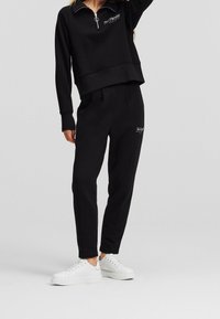 Person wearing black tracksuit with zippered collar and logo on chest and thigh, paired with white platform sneakers, standing on gray floor.