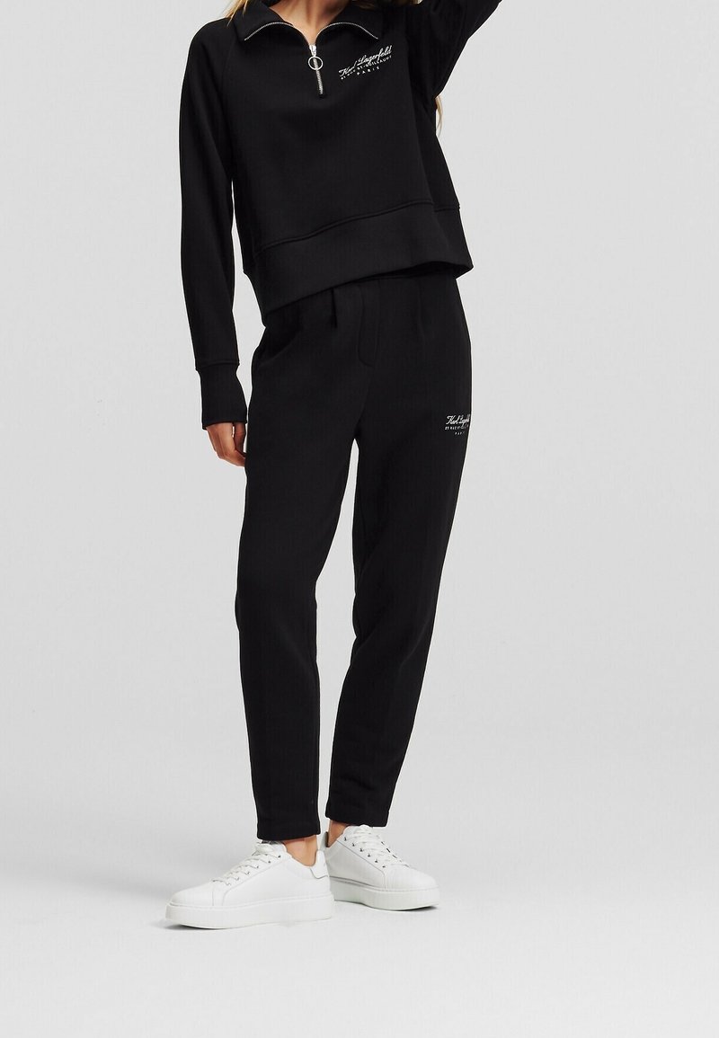 Person wearing black tracksuit with zippered collar and logo on chest and thigh, paired with white platform sneakers, standing on gray floor.