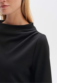 Top nero con tessuto testurizzato, scollatura alta, dettaglio drappeggiato sulla spalla e vestibilità ampia. Silhouette liscia con una sottile profondità.