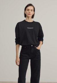 Juodas ilgomis rankovėmis marškinėliai su balta "Calvin Klein Jeans" logotipu ant krūtinės, derinami su aukštu liemeniu juodomis džinsomis. Lygus, kasdieniškas audinys.
