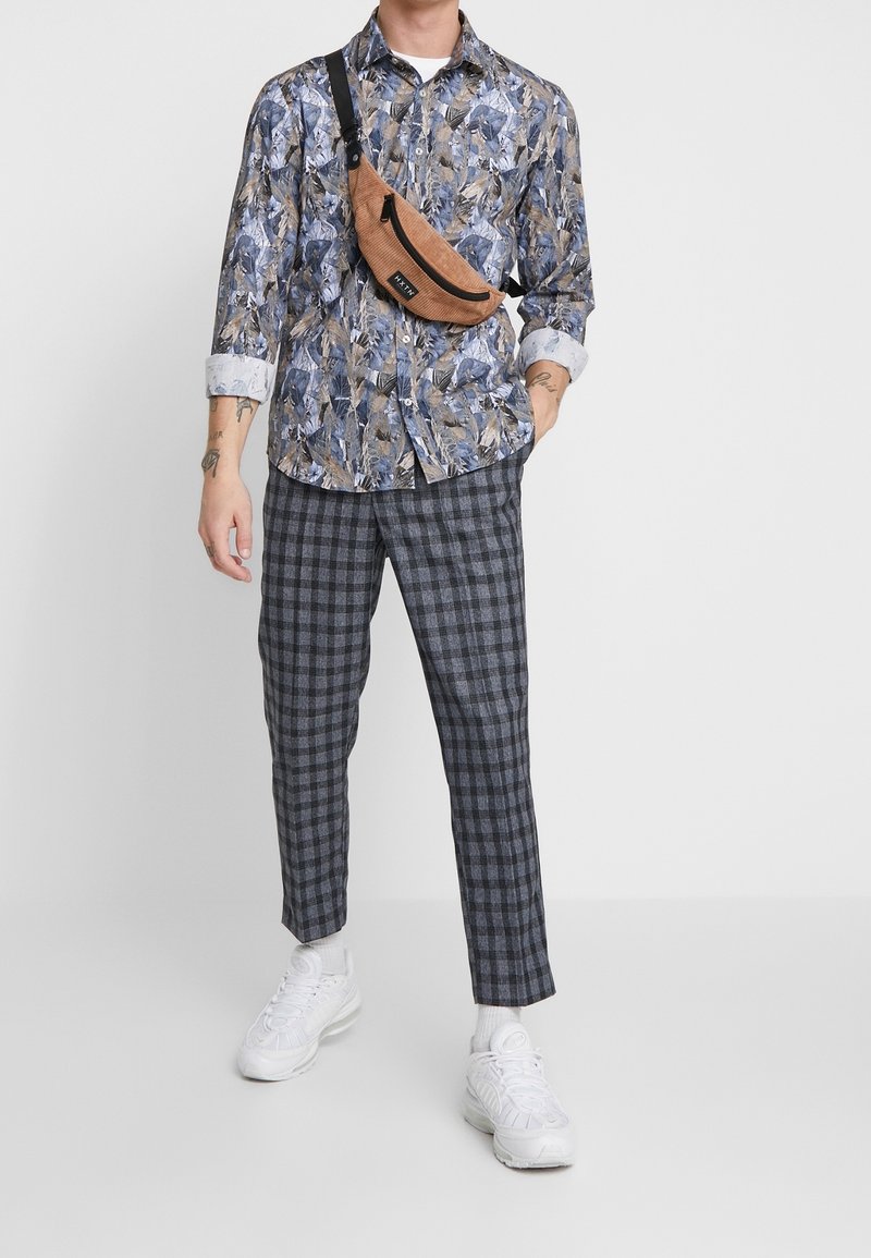 Blumenmusterhemden in Blau-, Braun- und Weißtönen, kombiniert mit karierten grauen Hosen. Braune Umhängetasche und weiße Sneakers.
