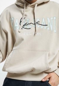 Sudadera beige con el texto "KALIKANI EST. 1989" y una línea cursiva negra superpuesta, combinada con jeans azul oscuro, con la mano descansando cerca del bolsillo.