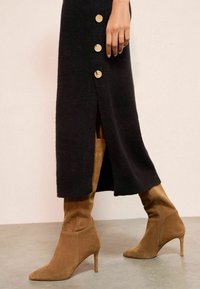 Rochie midi tricotată neagră cu butoane aurii și fante laterale, asortată cu cizme stiletto din suede bej, cu vârf ascuțit și fermoar.