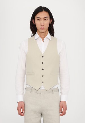 Beige vest med fem sorte knapper, to frontlommer, båret over en hvit skjorte. Lyse grå bukser kompletterer antrekket. Glatt stoffstruktur.