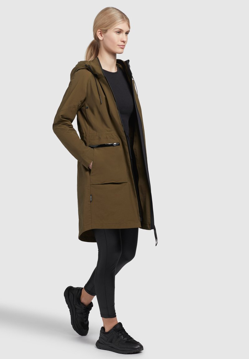 Olivgrüne, wasserdichte Jacke mit Kapuze, zwei Reißverschlusstaschen und verstellbaren Kordeln. Kombiniert mit schwarzen Leggings und Sneakers.