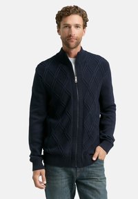 Homme portant un pull zippé bleu marine avec un motif en losanges, une chemise blanche en dessous, et un jean bleu, se tenant avec une main dans la poche.