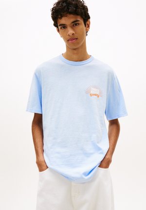 Ung mann som står med hendene i lommer, iført lyseblå T-skjorte med liten solstrålegrafikk på brystet og hvite bukser.