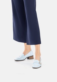 Mocassini in pelle azzurro chiaro con tacco in legno basso e dettaglio di fibbia dorata. Indossati con pantaloni culotte blu navy. Texture liscia, forma classica.