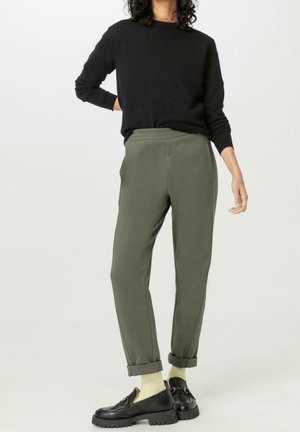 Person wearing dark long-sleeve top, olive green rolled-up pants, light-colored socks, and black chunky loafers, standing against plain background.