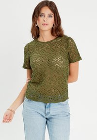 Haut en crochet vert à manches courtes, avec un motif floral et un design en maille ouverte. Texture légère et ourlet festonné. Assorti à un jean bleu.