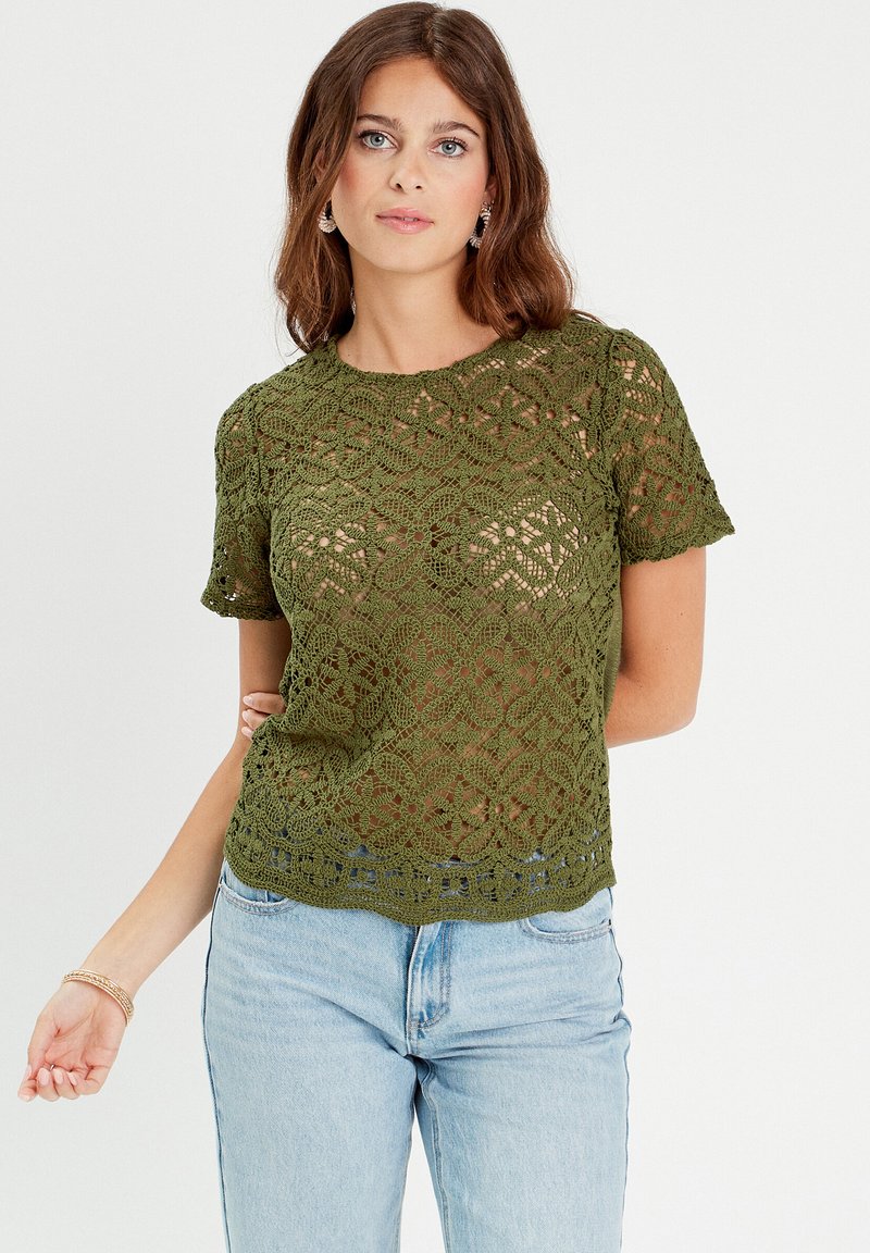 Haut en crochet vert à manches courtes, avec un motif floral et un design en maille ouverte. Texture légère et ourlet festonné. Assorti à un jean bleu.