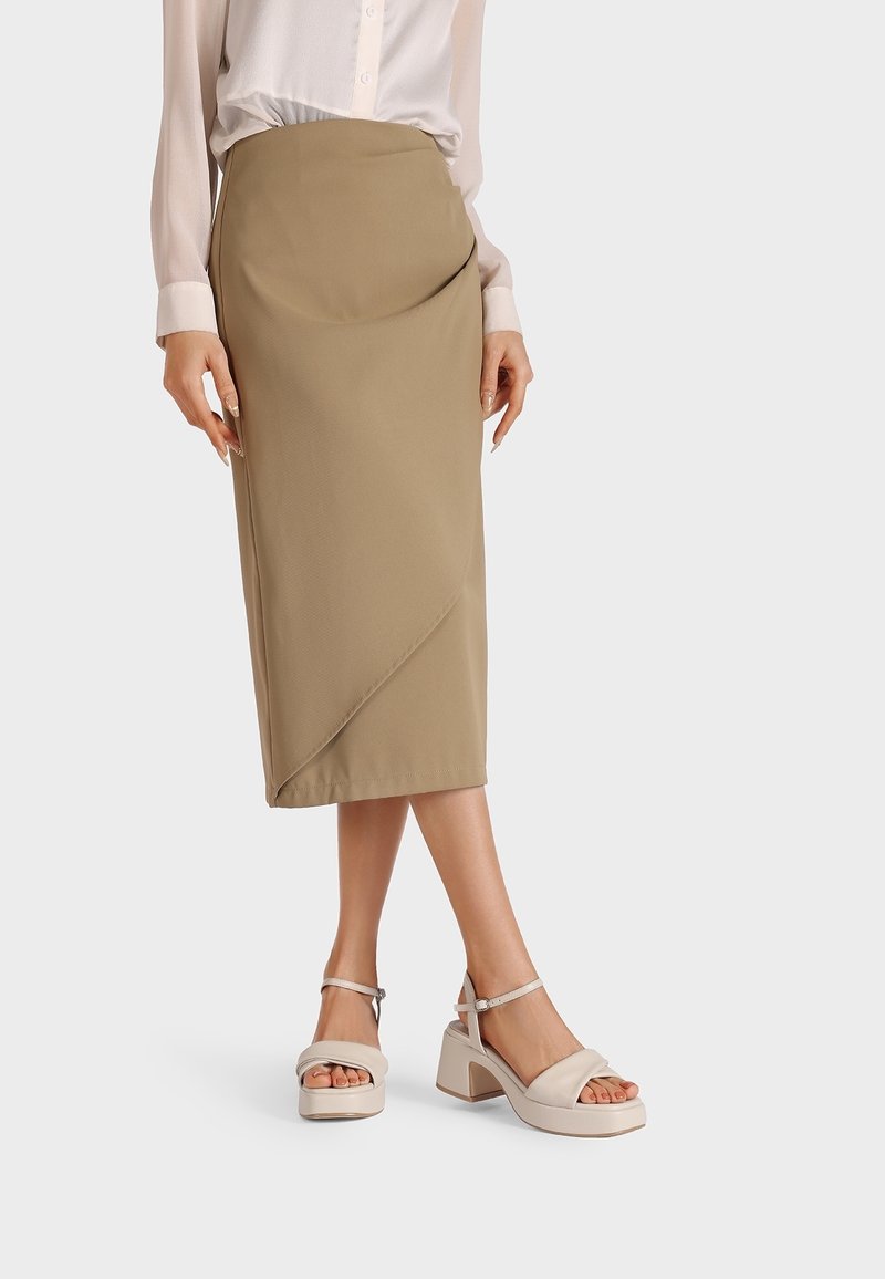 Femme portant une jupe midi beige asymétrique, un chemisier blanc et des sandales beiges à talons épais avec des lanières autour de la cheville, debout devant un fond blanc.
